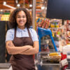 Pretty young smiling African-american female sales clerk with her arms crossed by chest looking at you by cash register during work