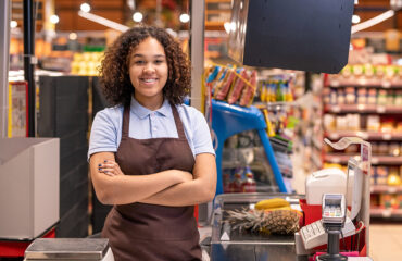 Pretty young smiling African-american female sales clerk with her arms crossed by chest looking at you by cash register during work