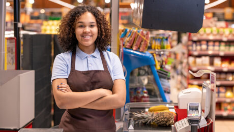 Pretty young smiling African-american female sales clerk with her arms crossed by chest looking at you by cash register during work