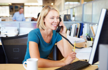 A smiling blonde woman in an office on the telephone